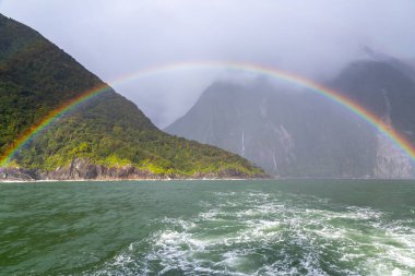 Yeni Zelanda 'nın Güney Adası' ndaki Milford Sound Fiordland Ulusal Parkı 'ndaki dağların önündeki suyun üzerinde renkli ve canlı bir gökkuşağının fotoğrafı.