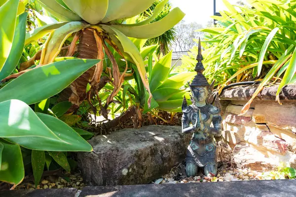 Küçük bir Tayland Buda heykelinin fotoğrafı. Bir yerleşim yerindeki yeşil bitkilerin arasında meditasyon pozu vermiş..