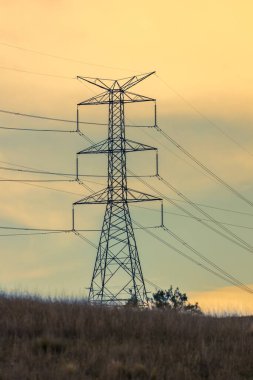 Avustralya 'nın turuncu gün batımında çimenli bir tepenin üzerindeki büyük bir elektrik iletim kulesinin fotoğrafı..