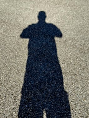 Arkalarından parlayan parlak güneş ışığı nedeniyle asfalt bir otoparkın zemininde duran büyük siyah bir gölgenin fotoğrafı..