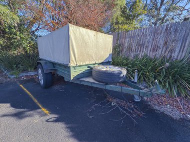 Eski, paslı, küçük, pis bir karavanın fotoğrafı. Kapalı bir tepsi ile kaplı. Küçük bir otoparkta, yerde ölü yapraklar ve ağaç dalları olan eski ahşap çitin yanında..