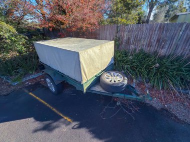 Eski, paslı, küçük, pis bir karavanın fotoğrafı. Kapalı bir tepsi ile kaplı. Küçük bir otoparkta, yerde ölü yapraklar ve ağaç dalları olan eski ahşap çitin yanında..