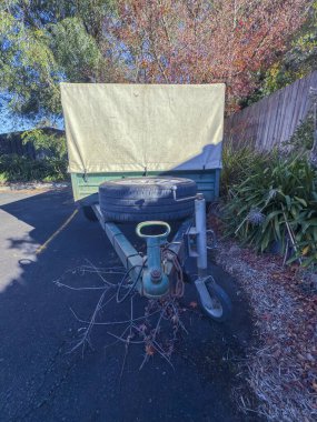 Eski, paslı, küçük, pis bir karavanın fotoğrafı. Kapalı bir tepsi ile kaplı. Küçük bir otoparkta, yerde ölü yapraklar ve ağaç dalları olan eski ahşap çitin yanında..