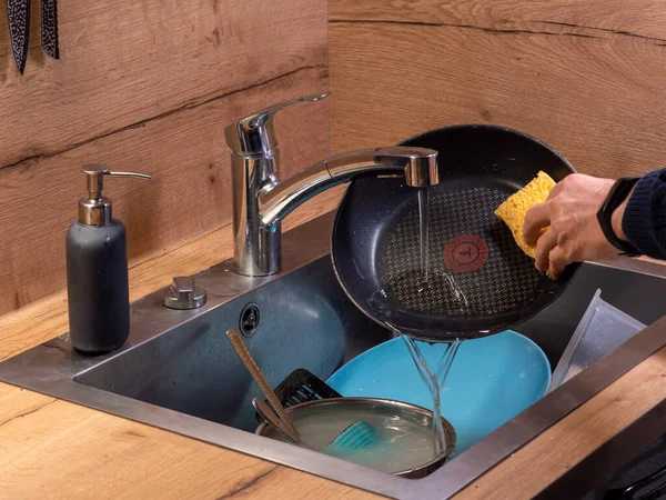 washing dishes by hand, a man washing pans, next to the sink there is a sink full of things to wash. Wooden background, chrome tap
