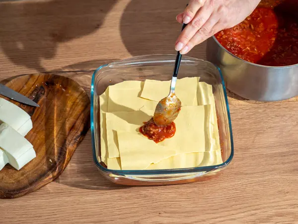 stock image Lasagna, an Italian dish, preparation, laying out tomato sauce with a metal spoon, next to it there is a metal pot with sauce, wooden countertop