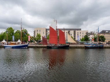 Caen, Fransa, 27 Haziran 2025. Denizcilik festivali sırasında rıhtımda kırmızı ve krema yelkenli tarihi yelkenli gemiler demirlemiştir. Renkli bayraklar ve modern kentsel binaların arkaplanı