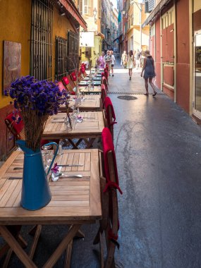 Güzel, Fransa, 8 Ağustos 2025. Nice 's Old Town' da dar bir sokakta restoran masaları. Kırmızı örtüler sandalyeleri kaplıyor ve masanın üzerinde dekoratif bir vazo çiçek duruyor. Turistler arka planda gezinirler.