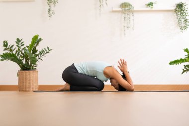 Fit and focused Asian woman practicing yoga in the comfort of her own home. Wearing comfortable clothing, she is performing various yoga poses in living room. Surrounded by natural light and peaceful 
