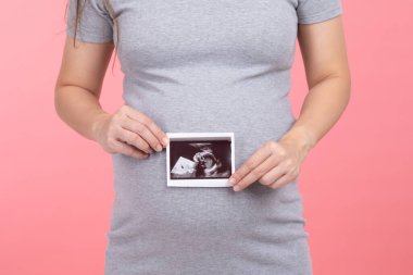 Mutlu hamile kadın, elinde ultrason görüntüsüyle ayakta duruyor pembe arka planda izole edilmiş aşkla koca göbeğini okşuyor, genç kadının hamileliği gelecekteki hayattan, annelikten ve hamilelikten zevk alıyor.