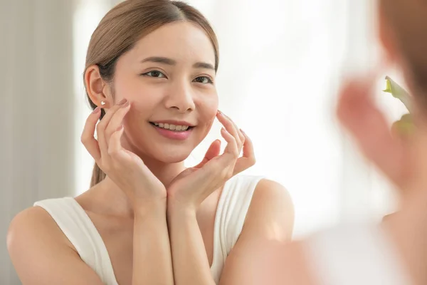 Back view of beautiful Asian young woman looking in the mirror after wake up preparing skin for make up. Healthy female looking her self in mirror smile and happiness with healthy skin. Self Care