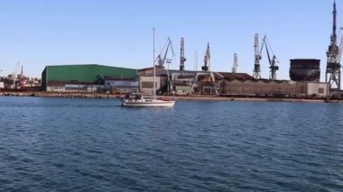 Açık bir yaz gününde, direkli bir yat denizde yüzüyor olacak. Ticari Ticaret Limanı, Turnalar ve Limanlar Arkaplanda.