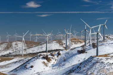 Wind turbines on a windmill farm for alternative energy production shown in the snow-covered Tehachapi mountains in Southern California. The San Gabriel Mountains are seen in the far distance.  clipart