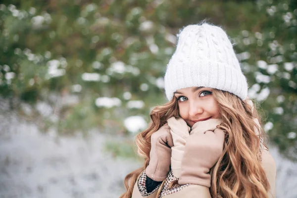 A sweet blonde girl against the backdrop of a winter snow-covered forest. Close-up photos. The concept of the new year, Christmas. A place for text. A girl in a white hat in a pine forest.