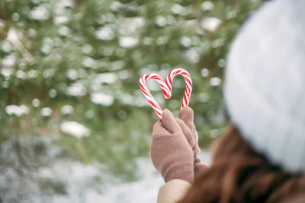 Close-up of women\'s hands holding candy canes in the form of heart. Small depth of field, selective focus. Sweets in the hands of a woman against the background of a snowy forest