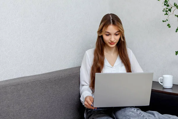 A young beautiful girl sits on the couch and holds a laptop in her arms. Online training. Communication in social networks. Interview for work. A place for text.