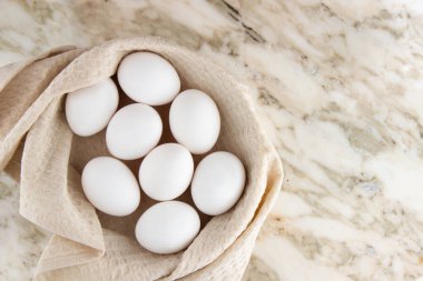 Beyaz tavuk yumurtaları bej bir waffle havlusuyla kaplı bir sepetin içinde mermer bir masanın üzerinde yatıyor. Paskalya bileşiminin üst görünümü, metin için boşluk