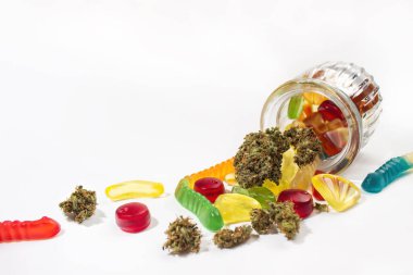 Kabartmalı cam kavanozdan çeşitli jelibonlar ve kurumuş tıbbi marihuana tomurcukları düştü. Beyaz bir arka planda. Bir sürü boş alan