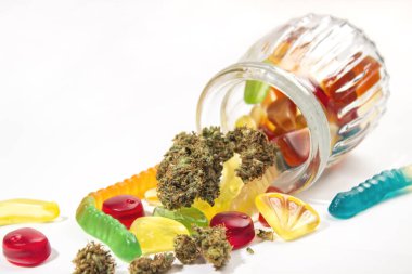Kabartmalı cam kavanozdan çeşitli jelibonlar ve kurumuş tıbbi marihuana tomurcukları düştü. Beyaz bir arka planda. Bir sürü boş alan
