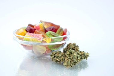 Assorted different gummy candies in a small glass bowl, near in the foreground several buds of dried medical marijuana, reflected on the glass clipart