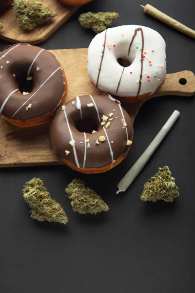 涂有巧克力和白冰的甜甜圈躺在医用大麻干芽的木板上 旁边有两个关节 在黑色背景上 — 图库照片