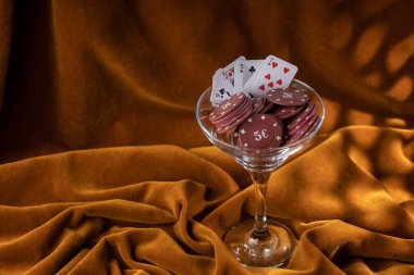 Kırmızı plastik poker çipleri, kahverengi kadife kumaşın üzerindeki cam margarita bardağında yatıyor. Bardağın kenarındaki iskambil kağıtlarının hayranıyım..  