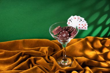 Kırmızı plastik poker çipleri, kahverengi kadife kumaşın üzerindeki cam margarita bardağında yatıyor. Bardağın kenarındaki küçük iskambil kağıtlarının hayranı, kokteyl garnitürü gibi