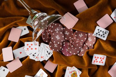 Cam margarita bardağından kahverengi kadife kumaşın üzerine kırmızı plastik poker fişleri düştü. Etrafta küçük oyun kartları var..