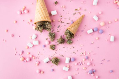 Kuru tıbbi marihuana tomurcukları pembe arka plandaki kremalı dondurma külahlarının üzerinde yatıyor. Etrafta şekerler ve marşmelovlar var. Alternatif tıbbi esrar tedavisi