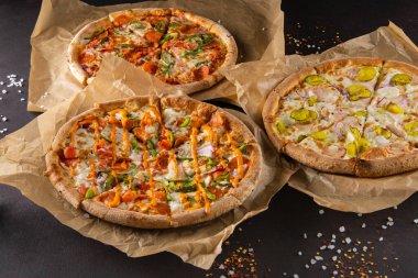 Üç güzel renkli, sulu farklı pizza siyah bir arkaplanda parşömen üzerinde yatıyor.