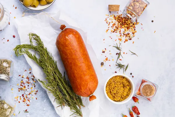 Baharat ve otlarla çevrili tütsülenmiş sosis mavi bir masanın üstünde, zeytin ve kumlu hardalın yanında.