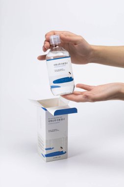 Huş suyu nemlendirici toner içeren minimalist bir kompozisyon. Kadın eli, yanında açık bir kutu olan şeffaf bir şişe tutuyor. Hafif arka plan saflık Yuvarlak Laboratuvarını vurgular