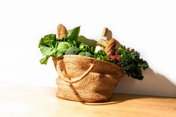 Shopping straw bag full of fresh leafy vegetables and a bottle of white wine. Strong sunny shadow. Healthy food ingredients shopping concept. Copy Space for a text.