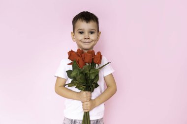 A boy in a white T-shirt, trousers on a pink background holds a bouquet of red roses in his hands. A gift for my mother, grandmother