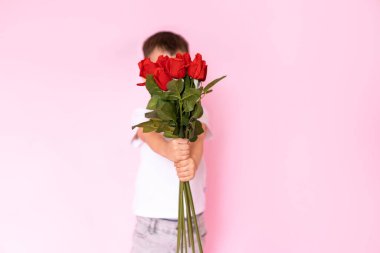 A boy in a white T-shirt, trousers on a pink background holds a bouquet of red roses in his hands and covers his face with them. A gift for my mother, grandmother