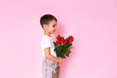 A boy in a white T-shirt, trousers on a pink background holds a bouquet of red roses in his hands and stands sideways and does not look at the camera. A gift for my mother, grandmother