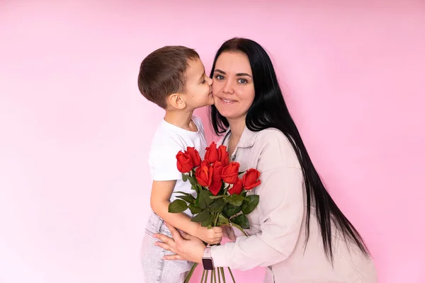 a gentle son kisses a happy mother and gives her a bouquet of roses, congratulating her on Mothers Day during the celebration of the holiday on a pink background