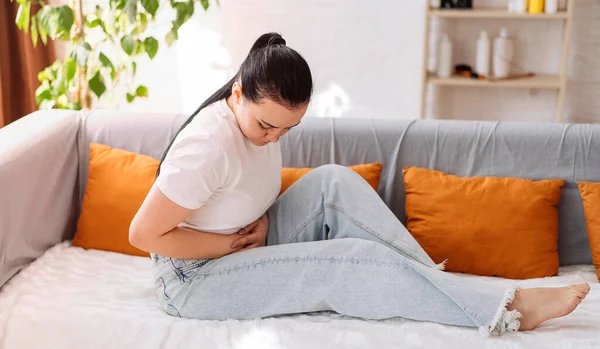 stock image A young woman suffering from severe abdominal pain while sitting on the couch at home. Close-up.