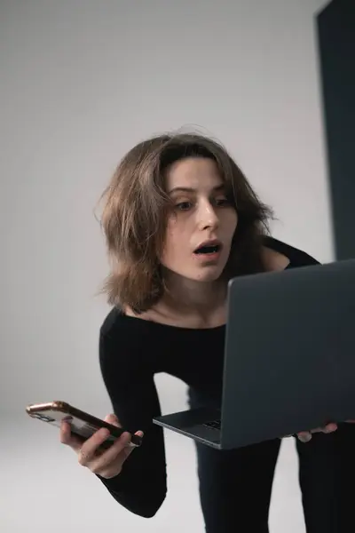 Şaşırmış bir kadın elinde telefonla dizüstü bilgisayara bakıyor..