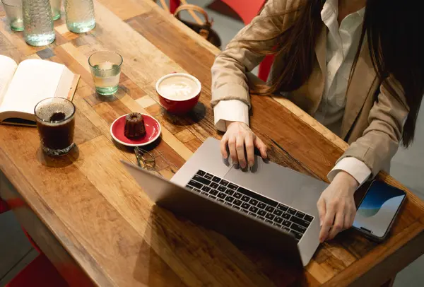 Dizüstü bilgisayar, kahve, pasta ve kadın eli olan bardaklar.