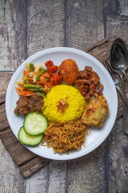 Indonesian traditional food, nasi kuning / yellow turmeric rice served with beef rendang, fried chicken, vegetables, spicy eggs, noodles, and tempeh