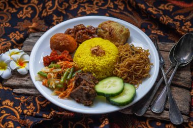 Indonesian traditional food, nasi kuning / yellow turmeric rice served with beef rendang, fried chicken, vegetables, spicy eggs, noodles, and tempeh