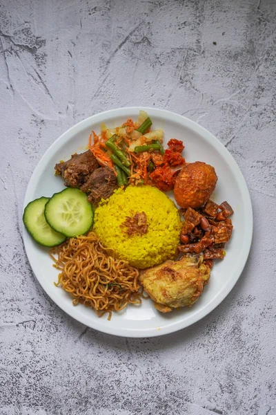 Indonesian traditional food, nasi kuning / yellow turmeric rice served with beef rendang, fried chicken, vegetables, spicy eggs, noodles, and tempeh