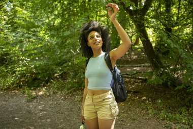 Genç bir kadın ormanda yürüyor, ağaçlara bakıyor.