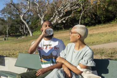 Çift bankta oturuyor ve su içiyor.