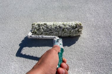İşçi evin çatısına akrilik beyaz boya sürüyor. Elinde silindir tutan koyu tenli bir işçi, evinin çatısını yalıtmak için beyaz boya sürüyor. Boşluk resmini kopyala.