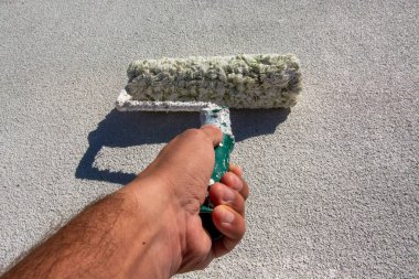 İşçi evin çatısına akrilik beyaz boya sürüyor. Elinde silindir tutan koyu tenli bir işçi, evinin çatısını yalıtmak için beyaz boya sürüyor. Boşluk resmini kopyala.
