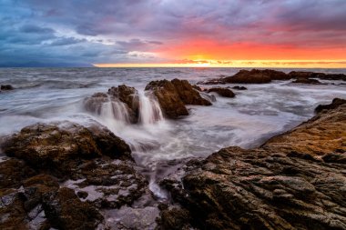 Kayalıklarda detayları olan ilham verici bir okyanus gün batımı ve kıyı yüksek çözünürlüğüne doğru yumuşak bir beyaz dalga geliyor.