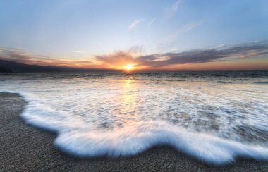 Renkli bir gün batımı gökyüzü ufkuyla kıyıya yuvarlanan bir dalganın geniş açılı yakın çekimi