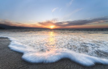 Renkli bir gün batımı gökyüzü ufkuyla kıyıya yuvarlanan bir dalganın geniş açılı yakın çekimi