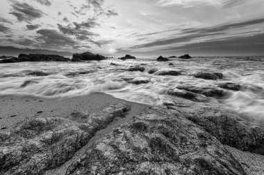 Deniz taşlarının üzerinde dalgaların akışına doğru yuvarlanan dalganın geniş açılı yakın çekimi dramatik bir siyah ve beyaz karşıtlık görüntüsüdür.